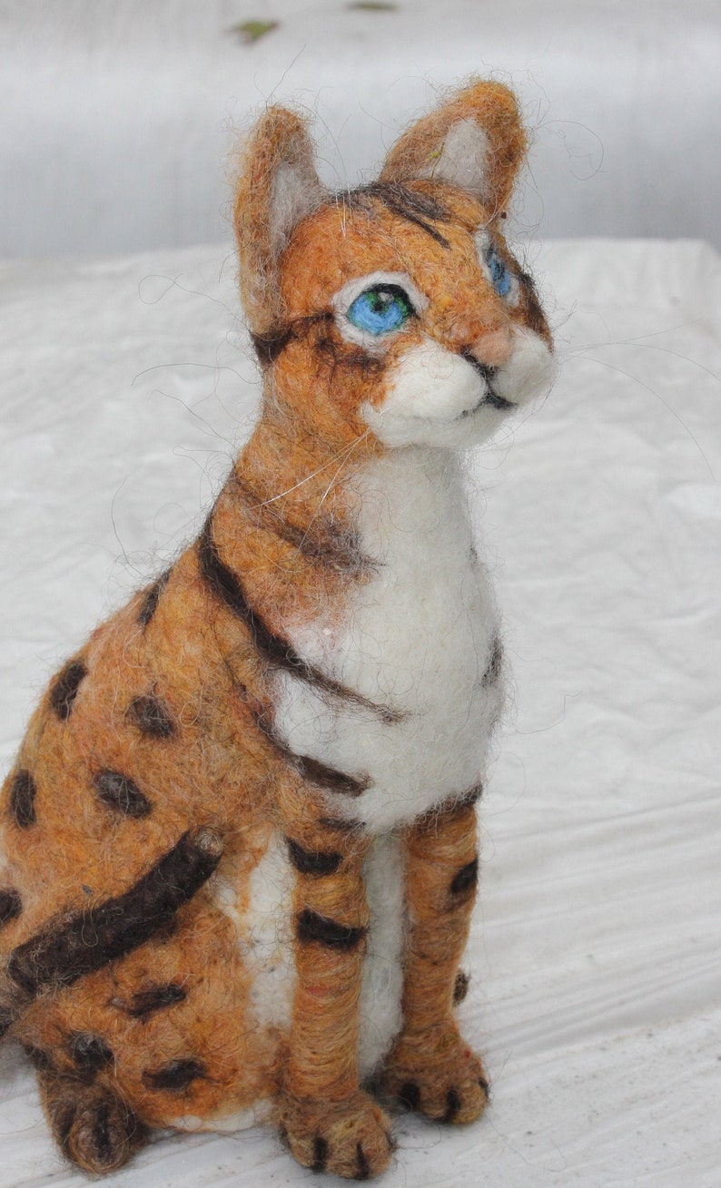 May include: A felted brown and white tabby cat with blue eyes. The cat is sitting on a white surface and looking to the right.