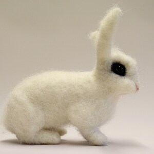 May include: A white, needle-felted rabbit with black eyes. The rabbit is sitting on a white surface.
