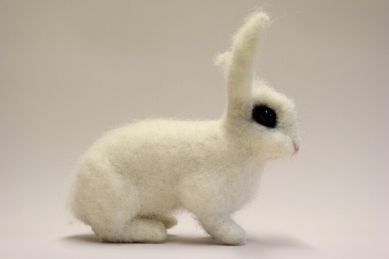 May include: A white, needle-felted rabbit with black eyes. The rabbit is sitting on a white surface.