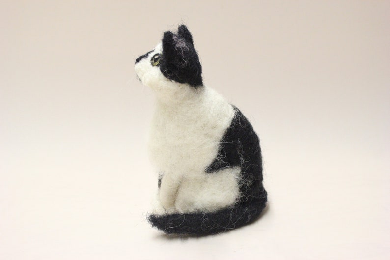 May include: A black and white felted cat figurine. The cat is sitting with its head turned to the side and its tail curled around its body.