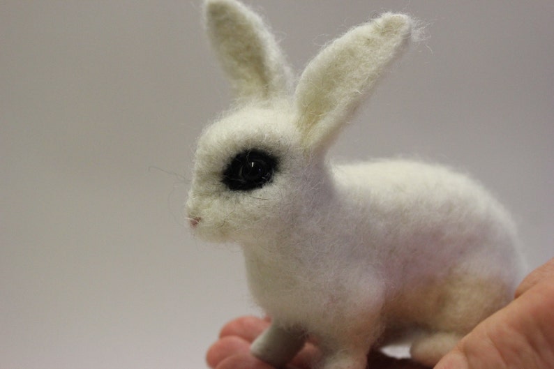 May include: A white, needle-felted rabbit with black eyes. The rabbit is sitting on a hand.