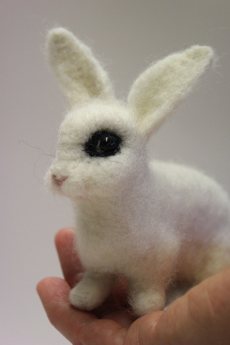 May include: A white, needle-felted rabbit with black eyes. The rabbit is sitting in a person's hand.