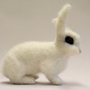May include: A white, fluffy rabbit with black eyes is sitting on a white surface. The rabbit is facing to the right and has its ears perked up.