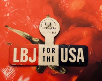 May include: A vintage political campaign button with the text "LBJ for the USA" in white letters on a blue background. The button is attached to a white tag with a small, round sticker that says "United Duck Club of Chicago" and has a number on it.