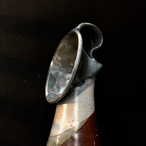 May include: A ceramic vase with a unique, twisted design. The vase is glazed in shades of brown, white, and gray, with a speckled finish. It has a wide, open mouth and a single handle.