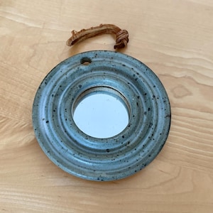 May include: A small, round, ceramic mirror with a speckled blue-grey finish. The mirror has a central reflective surface and a loop of twisted, brown material for hanging. The mirror is approximately 10 cm in diameter.
