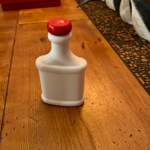 May include: A white ceramic bottle with a red plastic lid. The bottle is empty and has a rounded rectangular shape.