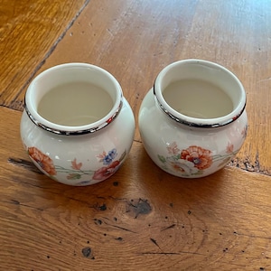 May include: Two small, round, white ceramic vases with a floral design featuring red, orange, and blue flowers. Each vase has a silver rim and sits on a wooden surface. The vases are empty.