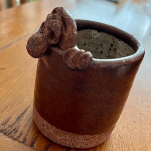 May include: A brown ceramic mug with a textured surface and a sculpted animal head handle. The mug has a speckled glaze and a brown rim.
