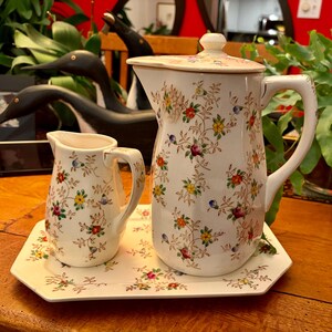 May include: A white ceramic pitcher with a lid, a creamer, and a tray, all adorned with a floral pattern in red, yellow, blue, and green. The pitcher has a large handle and a spout. The creamer has a small handle. The tray is octagonal.
