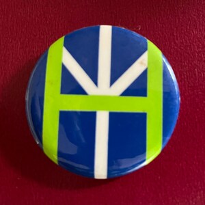 May include: A round button with a blue background and a white and green design. The design features a white cross with green lines extending from the centre.