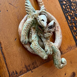 May include: A ceramic dragon figurine with green and brown scales, holding a small, white, ceramic bird in its claws. The dragon is curled around a circular, brown, ceramic base. The figurine is hanging from a rope with yellow beads.