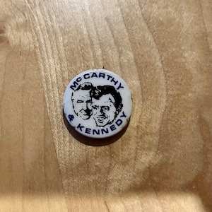 May include: A white button with a black and white image of two men's faces. The text "MCCARTHY & KENNEDY" is printed in blue around the edge of the button.