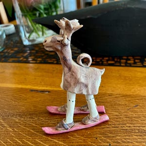 Puede incluir: Una figurita de cerámica de un reno con un abrigo marrón y blanco y esquiando en esquís rosas.