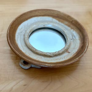 May include: A small, round, ceramic mirror with a brown and cream-colored frame. The mirror is set in a shallow dish with a decorative, textured border. The base has two small, curved feet for support. The mirror reflects the surrounding environment.