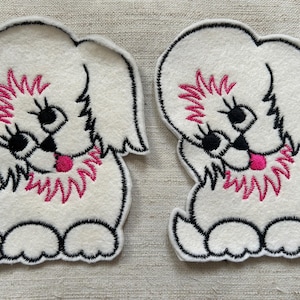 May include: Two white embroidered patches of a cartoon dog. The dog has black eyes, a pink tongue, and a pink collar. The patches are outlined in black.