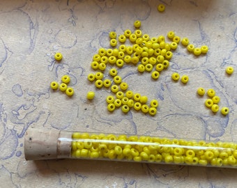 May include: A glass tube filled with small, bright yellow beads. The beads are scattered on a light brown surface with blue and white marbling.