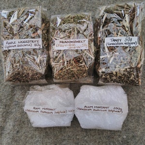 May include: Five packages of natural dyeing supplies are arranged in a top-down view. Three bags hold dried herbs: "Purple Loosestrife," "Meadowsweet," and "Tansy," each at 50g. Two packages of "Alum Mordant" (Potassium Aluminium Sulphate) are also present, weighing 100g and 200g.
