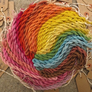 Könnte beinhalten: Eine Sammlung von 20 handgefärbten Stickgarnen, die in einem Regenbogenmuster angeordnet sind. Das Garn ist spiralförmig aufgewickelt, wobei jedes Knäuel mit einem Farbnamen beschriftet ist. Die Farben umfassen staubiges Orange, Persimons Orange, blasses Waidblau, Flechtenviolett und Konfetti.