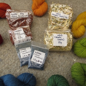 May include: A selection of natural dyeing ingredients and yarn. The image displays bags of Madder, Weld, and Woad, with handwritten labels showing weights in grams. Various skeins of yarn in shades of orange, yellow, green, and blue are also visible.