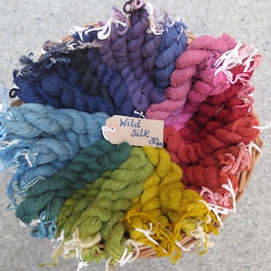 May include: A wicker basket filled with skeins of wild silk yarn in a rainbow of colors, including deep blue, teal, green, yellow, orange, red, pink, and purple. A tag reads "Wild Silk 30m".