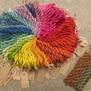 Może przedstawiać: Tęczowa kolekcja szpul z nicią do haftu ułożonych w kółko z brązowymi papierowymi etykietami z nazwami kolorów i numerami. Kolory nici wahają się od ciemnoniebieskiego do czerwonego.