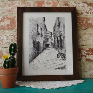 May include: A black and white ink drawing of a narrow street with buildings on either side. A bicycle is parked in the street. The drawing is framed in a brown wooden frame.