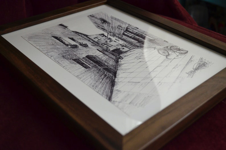 May include: A framed black and white pen and ink drawing of a narrow alleyway with buildings on either side. The drawing is in a dark brown wood frame.
