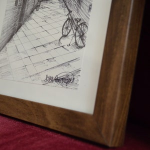 May include: A framed black and white ink drawing of a bicycle parked in a cobblestone alleyway. The drawing is signed "Jen Armstrong".