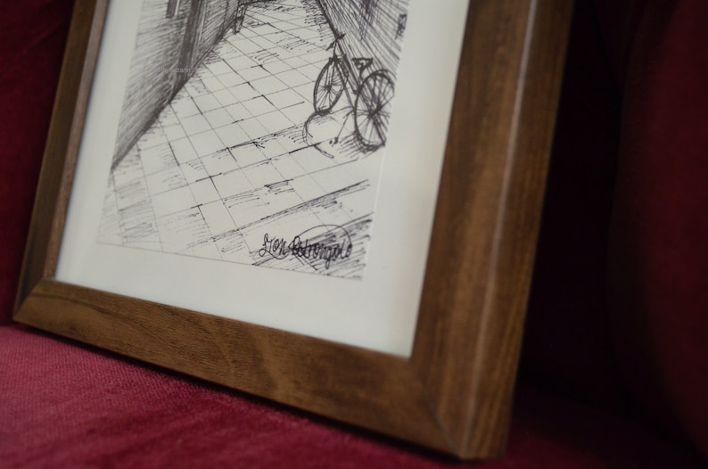 May include: A framed black and white ink drawing of a bicycle parked in a cobblestone alleyway. The drawing is signed "Jen Armstrong".