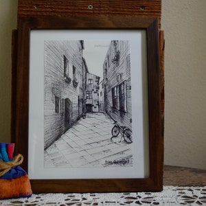 May include: A black and white ink drawing of a narrow street with buildings on either side. A bicycle is parked in the street. The drawing is framed in a brown wooden frame.