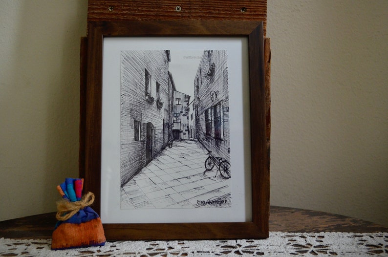 May include: A black and white ink drawing of a narrow street with buildings on either side. A bicycle is parked in the street. The drawing is framed in a brown wooden frame.