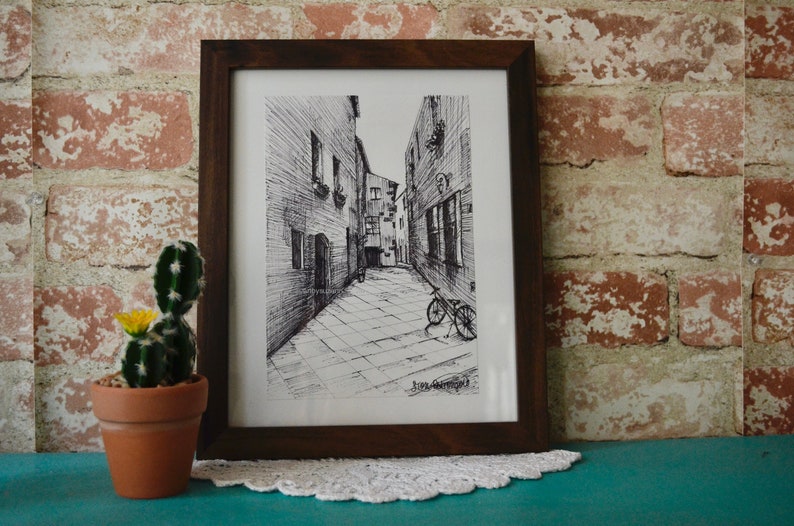 May include: A black and white ink drawing of a narrow street with buildings on either side. A bicycle is parked in the street. The drawing is framed in a brown wooden frame.