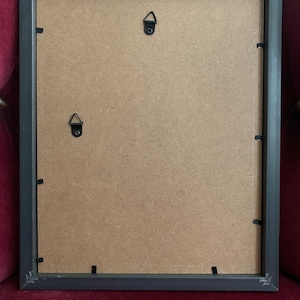 May include: Black picture frame with a brown cardboard backing. The frame has two metal hanging hooks on the back.
