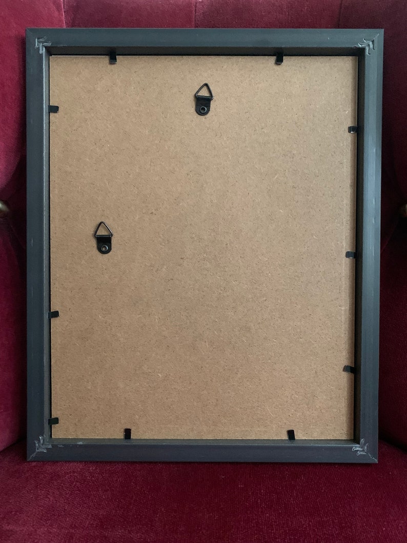 May include: Black picture frame with a brown cardboard backing. The frame has two metal hanging hooks on the back.