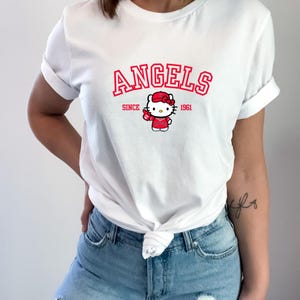 Puede incluir: Camiseta blanca con la palabra "ANGELS" en rojo sobre un dibujo de Hello Kitty con uniforme de b&eacute;isbol. El texto "SINCE 1961" tambi&eacute;n es rojo. La camiseta es de estilo casual.