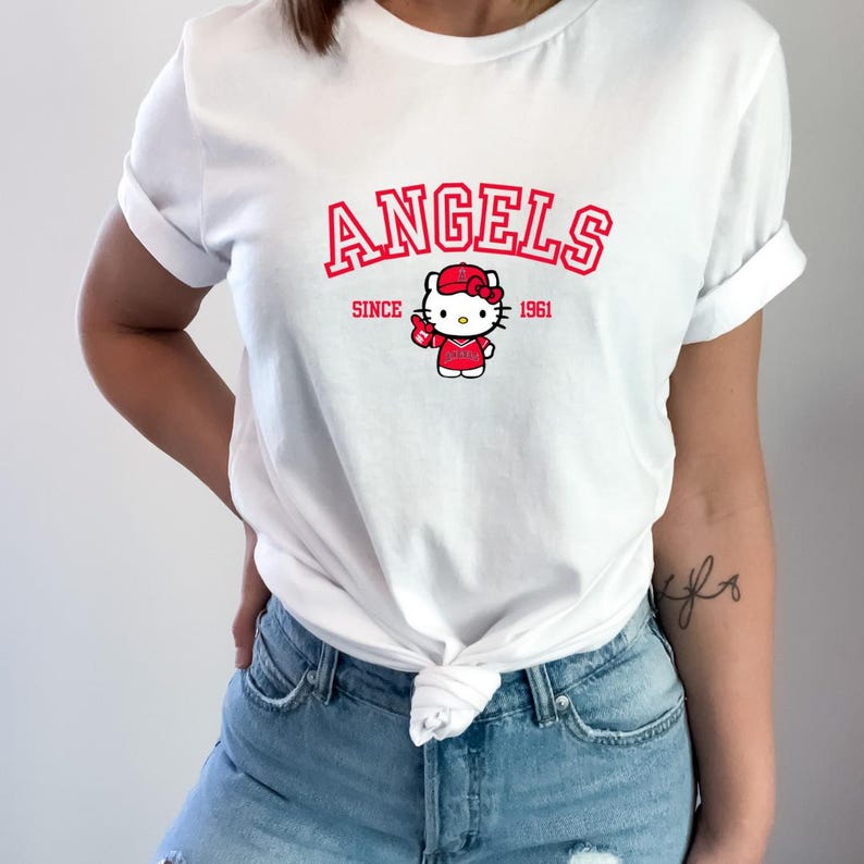 Puede incluir: Camiseta blanca con la palabra "ANGELS" en rojo sobre un dibujo de Hello Kitty con uniforme de b&eacute;isbol. El texto "SINCE 1961" tambi&eacute;n es rojo. La camiseta es de estilo casual.
