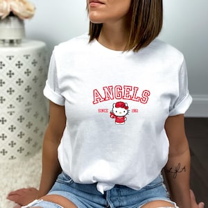 Puede incluir: Camiseta blanca con la palabra "ANGELS" en rojo, con "SINCE 1961" debajo. Un personaje de dibujos animados con un atuendo rojo y un lazo est&aacute; centrado debajo del texto. La camiseta es de estilo casual y de manga corta.
