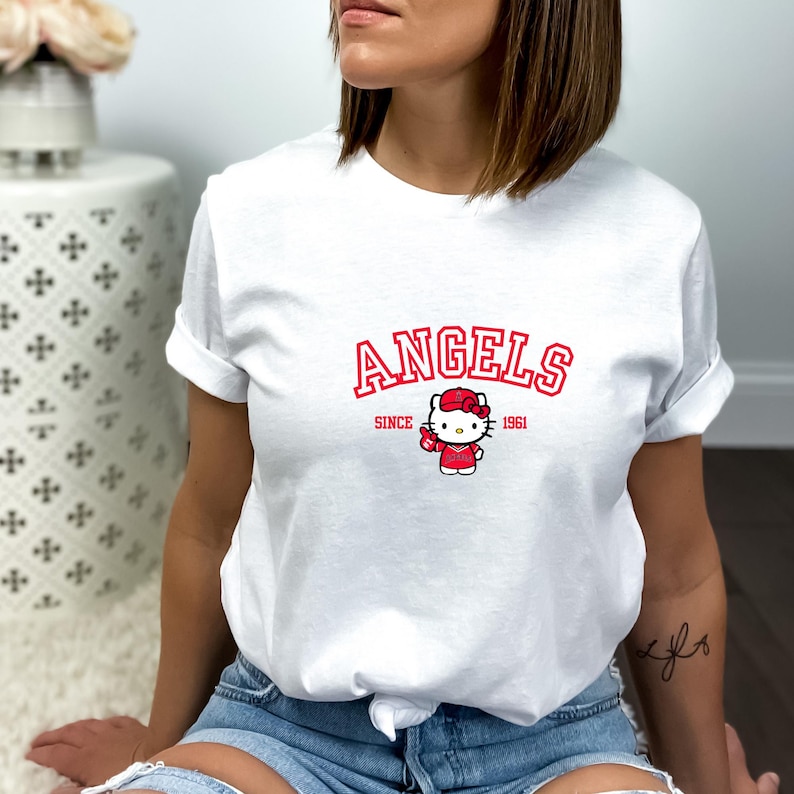 Puede incluir: Camiseta blanca con la palabra "ANGELS" en rojo, con "SINCE 1961" debajo. Un personaje de dibujos animados con un atuendo rojo y un lazo est&aacute; centrado debajo del texto. La camiseta es de estilo casual y de manga corta.