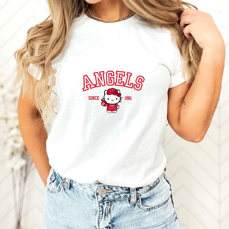 Puede incluir: Camiseta blanca con la palabra "ANGELS" en rojo, arqueada sobre un dibujo de Hello Kitty con atuendo y sombrero rojos. El texto "SINCE 1961" est&aacute; debajo. Una prenda informal para el d&iacute;a a d&iacute;a.