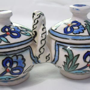 May include: A ceramic salt and pepper set with a handle, featuring a white base and lids. The set is decorated with blue floral patterns, green leaves, and a blue border. The handle has a decorative design.