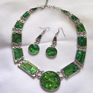 May include: A matching jewelry set featuring a necklace and earrings. The necklace and earrings have a silver-tone metal setting with vibrant green, rectangular and circular iridescent inlays. The necklace has a large circular pendant.