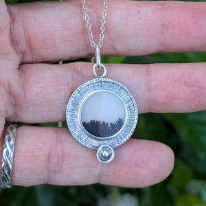 May include: A silver pendant necklace featuring a round, textured pendant with a black and white landscape design. The pendant hangs from a delicate silver chain. A small, round silver charm is below the pendant.