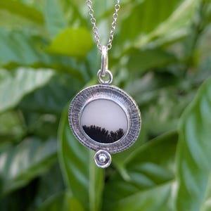 May include: A silver pendant necklace featuring a round, textured pendant with a black and white landscape design. The pendant hangs from a delicate silver chain. The background is blurred green foliage.