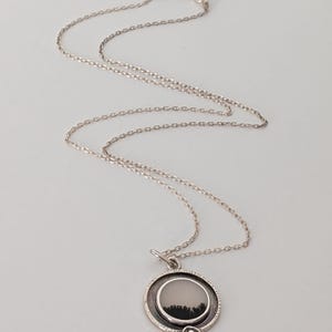 May include: A silver necklace with a circular pendant. The pendant features a landscape design with a black forest silhouette against a light background. The chain is delicate and hangs on a light background.
