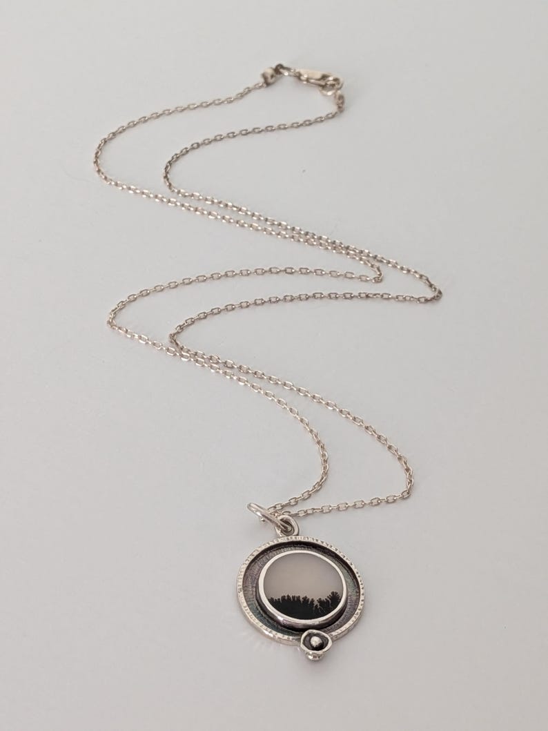 May include: A silver necklace with a circular pendant. The pendant features a landscape design with a black forest silhouette against a light background. The chain is delicate and hangs on a light background.