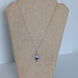 May include: A silver necklace with a circular pendant featuring a black and white landscape scene. The pendant is displayed on a tan jewelry stand against a white background. The necklace chain is delicate.