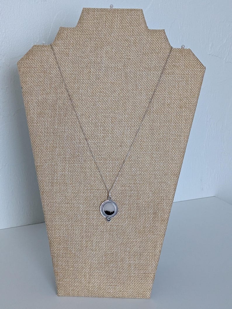 May include: A silver necklace with a circular pendant featuring a black and white landscape scene. The pendant is displayed on a tan jewelry stand against a white background. The necklace chain is delicate.