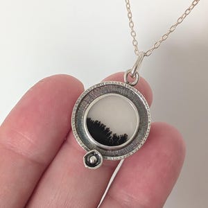 May include: A silver pendant necklace featuring a circular pendant with a white background and a black, tree-like design. The pendant is attached to a delicate silver chain. The necklace is held in a hand.