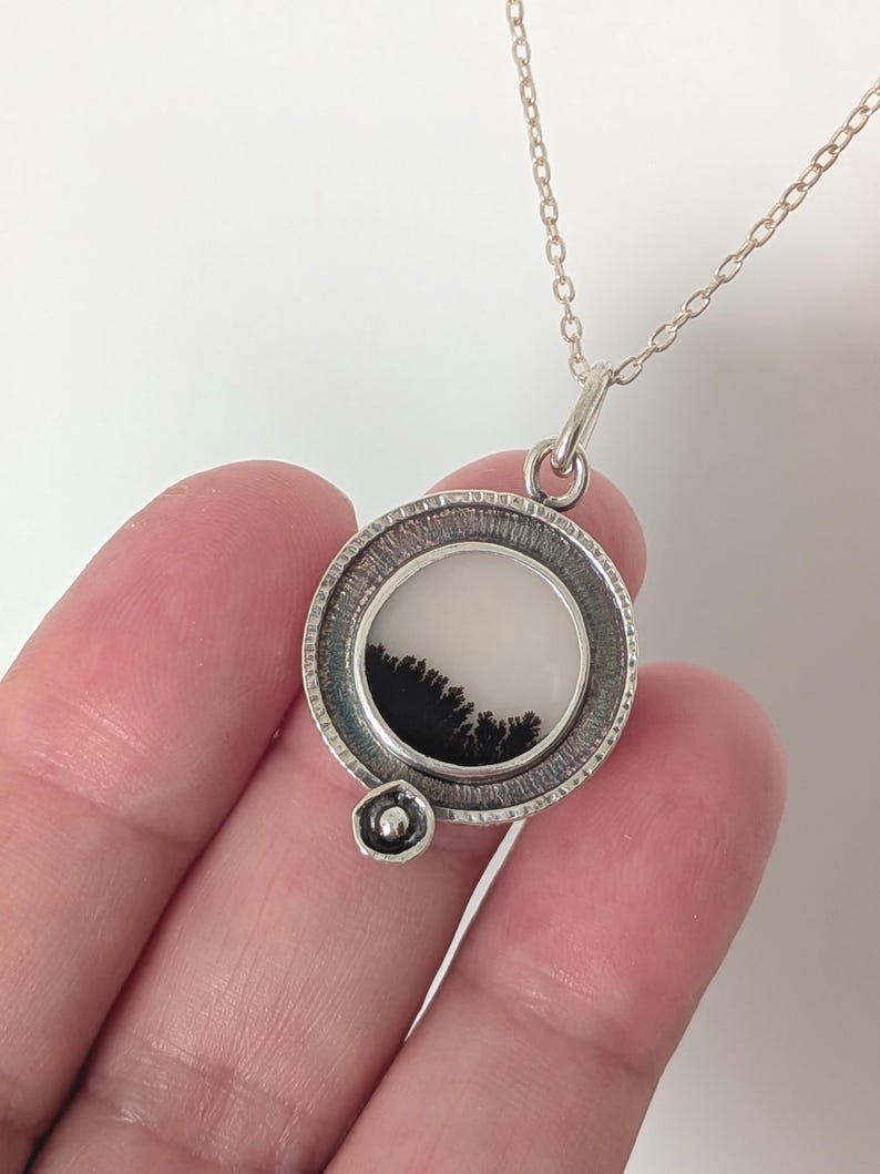 May include: A silver pendant necklace featuring a circular pendant with a white background and a black, tree-like design. The pendant is attached to a delicate silver chain. The necklace is held in a hand.
